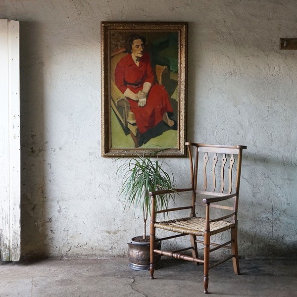 An antique wooden chair in front of a roughly plastered wall with and antique portrait hanging on it.
