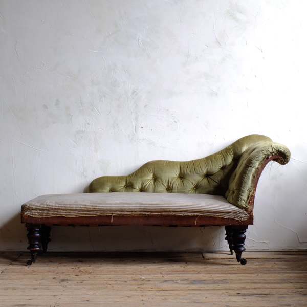 A mid-19th century scroll-arm chaise longue against a grey plastered wall.