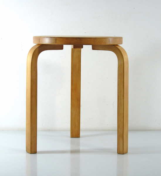 A simple plywood stool in a sparse room with white washed walls.