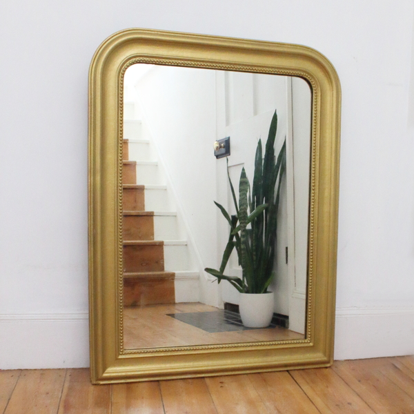 A large gold framed mirror reflecting bare wood stairs and a potted plant