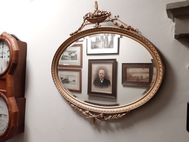 An oval-shaped ornate gilt mirror hung on a gallery wall.