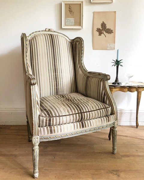 A wingback French antique armchair with striped upholstery next to a side table with muted tone art work on the wall behind.